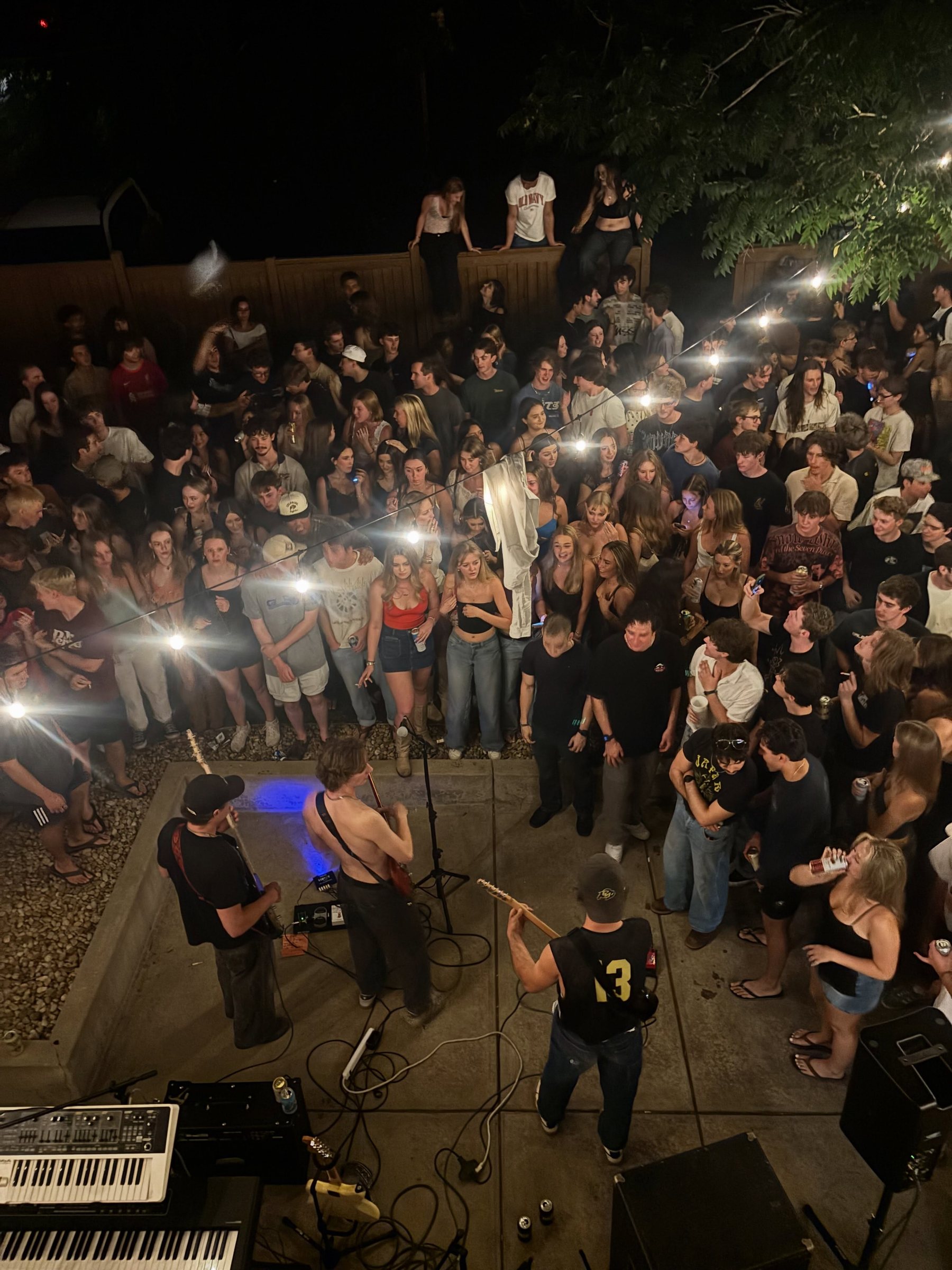Overhead view of a packed backyard show with a couple hundred people shoulder to shoulder, fans sitting on the fence to see over the crowd, and No Parachute playing on the patio below